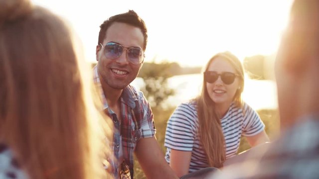 Close up view of beautiful young girl and boy in sunglasses talking to their friends while having a picnic at the riverbank, laughing heartily in a bright sunlight. Bright sunset, having leisure time.