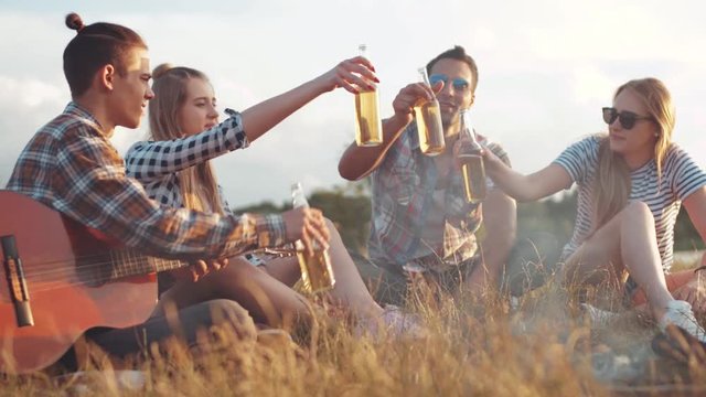 Close up view of young people gathered around the campfire, talking and toasting with the bottles of cyber. One of the friends holds the guitar. Being happy, young and free. Summertime, bright sunset.