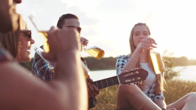 Young happy friends in casual wear toasting with bottles of beer while being at picnic, laughing happily. Beautiful nature, summer season. True emotions, positive mood.