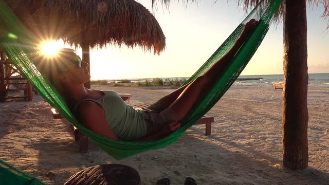 SLOW MOTION CLOSE UP: Pretty girl on summer vacations lying in a hammock swinging under a sunshade on stunning sandy beach at golden light sunset. Attractive woman with sunglasses sleeping at seashore