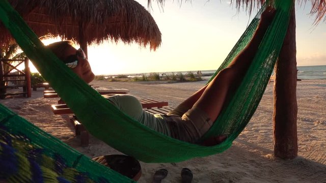 SLOW MOTION CLOSE UP Pretty girl on summer vacations lying in hammock swinging under a sunshade on beautiful sandy beach at golden light sunrise. Attractive woman with sunglasses sleeping at seashore