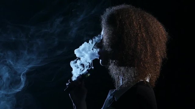Girl of african american appearance smokes an electronic cigarette. Black background. Side view. Silhouette. Slow motion