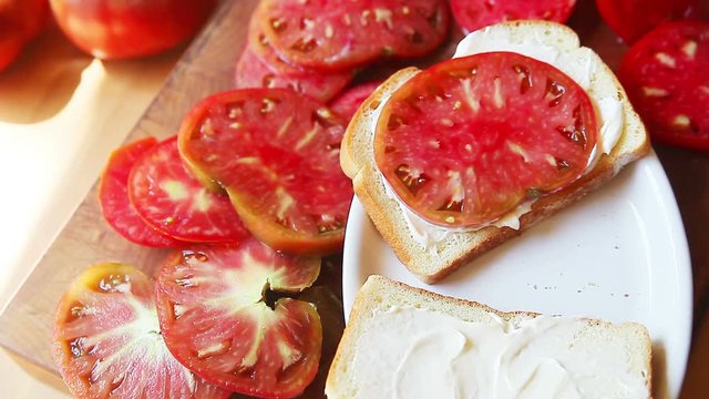 A senior man puts heirloom tomato slices on bread spread with mayonnaise with several slices alongside