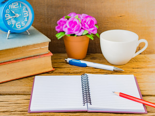 Office accessories including note book, red pencil, alarm clock and flower on wooden background. Education and business concept.