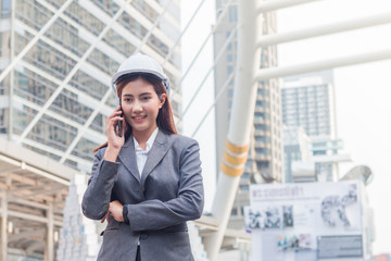 Portrait of beautiful young engineer woman wear a white safety helmet. She are smile and talking on smartphone with commitment to success with building background