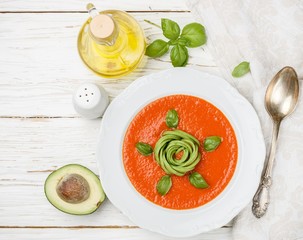 Delicious tomato soup with avocado, fresh Basil, olive oil and spices in a white plate on the table. The Mediterranean cuisine. Top view. Selective focus