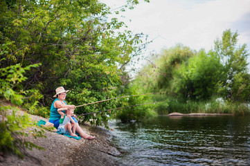 Fototapeta na wymiar Grandmother and grandson in green T-shirts sit on the river bank and fish. Grandmother with her grandson in straw hats is fishing in the summer