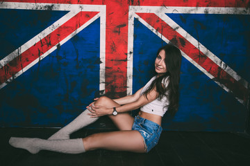 Красивая девушка с черными волосами сидит на фоне британского флага в коротких джинсовых шортах, гольфах и белой майке. Съемка в студии