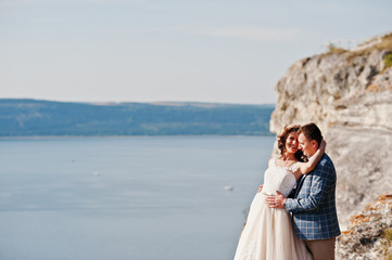 Fototapeta na wymiar Fantastic wedding couple standing on the edge of rocky precipice with a perfect view of lake on the background.