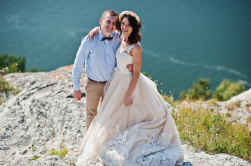 Fantastic wedding couple standing on the edge of rocky precipice with a perfect view of lake on the background.