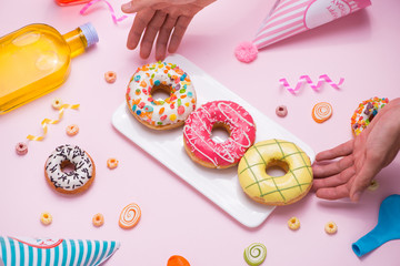 Party. Hand holding plate of colourful sugary round glazed donuts and bottles of drinks on pink background.
