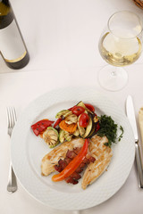Chicken with bacon and grilled vegetables arranged on a plate, Wine bottle and wineglass in background, Traditional dish in elegant setting, Selective focus with soft light