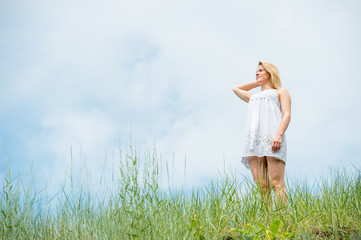 Naklejka na ściany i meble Beautiful girl in short white dress, posing on a meadow on warm sunny day. Sky and grass background