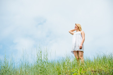 Beautiful woman in short white dress, posing and smiling on a meadow on warm sunny day. Blue sky and grass background.