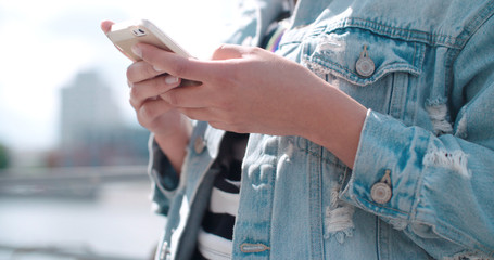 Unrecognizable portrait of young woman in denim jacket typing on phone in a city park during sunny day.