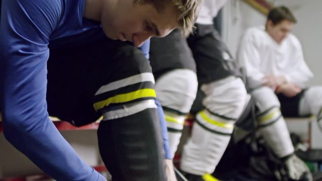 Tilt up of concentrated young man sitting on bench in changing room and tying shoelaces on his ice skates; teammates getting ready in background