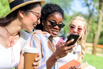 Obraz premium smiling multiethnic girls using smartphone and drinking coffee from paper cup in park