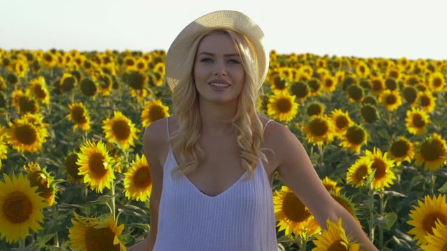 Young beautiful blonde woman standing in sunflower field. Sunset background. Sexy sensual portrait of girl in straw hat and white summer dress. Slow motion.