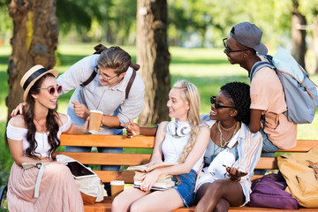 young multiethnic students holding books and digital devices while interacting in park