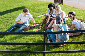 smiling young multiethnic students reading books while resting on grass in park