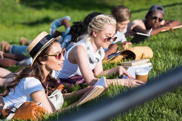 beautiful multiethnic girls reading books while lying on grass and studying in park