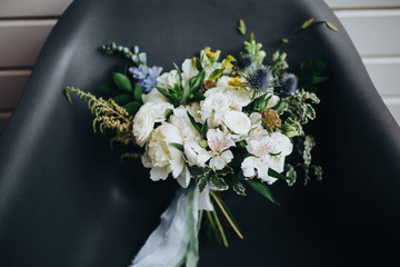 Wedding bridal bouquet of white flowers and greenery with ribbons standing on a black chair on background of white wooden wall