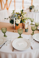 In the wedding banquet area there are wooden tables with tablecloths, on tables there are compositions of flowers and greens, candles, cutlery, on plates lie napkins with name cards