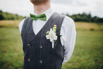 groom in a waistcoat and bow tie standing in a field, on the waistcoat a buttonhole of flowers and...