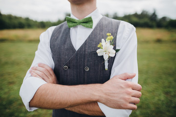 groom in a waistcoat and bow tie standing in a field, on the waistcoat a buttonhole of flowers and...