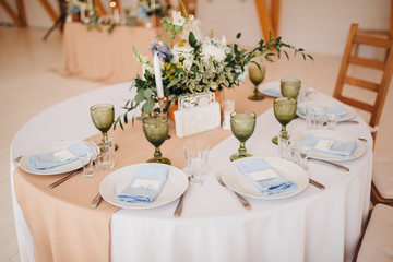 In the wedding banquet area there are wooden tables with tablecloths, on tables there are compositions of flowers and greens, candles, cutlery, on plates lie napkins with name cards