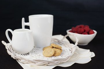 Cup of coffee, cookies a jug of milk on a straw tray. A bowl with raspberries. Black background