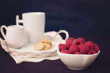 A bowl with raspberries. Cup of coffee, cookies a jug of milk on a straw tray. Black background. Vintage tinted