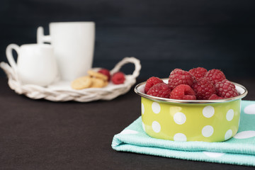 Green bowl with polka dot full with raspberries. Cup of coffee, cookies a jug of milk on a straw...
