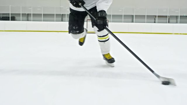 Tracking low-section shot of unrecognizable hockey player in white uniform dribbling puck in ice rink during practice
