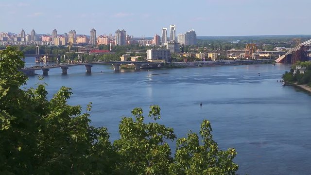 Capital of Ukraine - Kyiv city. Top view of cityscape, blue Dnipro, traffic on bridge over river and beautiful huge green trees in foreground. Real time full hd video footage.