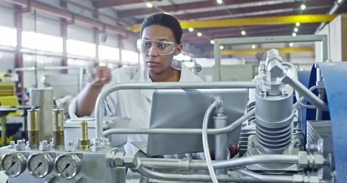 Slowmo of African female engineer in lab coat and protective eyeglasses typing on laptop while operating factory machine