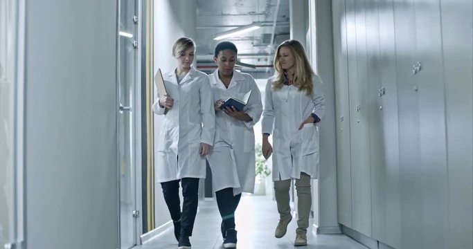 Slowmo of African female chief engineer in lab coat walking with two colleagues through corridor, holding organizer and talking to colleagues