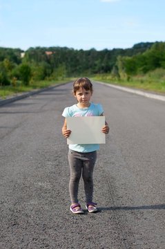 Sad little girl is standing on the road and is holding a piece of paper.