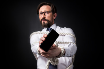 scared mature businessman with tied hands holding smartphone with blank screen isolated on black