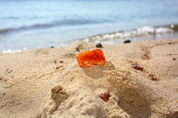 Amber. A red piece of Baltic amber on a sandy beach against a background of sea waves. Natural mineral amber in the sand lit by the sun. Frozen resin as an ornament in jewelry production.