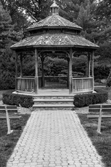 Front of gazebo with wood and steel benches in front. Outdoor gazebo with shrubs and grass. Shady retreat in backyard. Outdoor rotunda summerhouse. Outside platform.