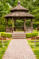 Front of gazebo with wood and steel benches in front. Outdoor gazebo with shrubs and grass. Shady retreat in backyard. Outdoor rotunda summerhouse. Outside platform. - obrazy, fototapety, plakaty