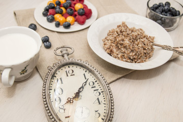 Buckwheat breakfast meal with berries on white plate, milk, blueberry, golden berry, physalis, raspberry, clock with 8 o'clock time 