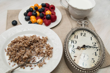 Buckwheat breakfast meal with berries on white plate, milk, blueberry, golden berry, physalis, raspberry, clock with 8 o'clock time 