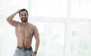 Fototapeta na wymiar Sexy fashion portrait of a hot bearded male model in stylish jeans with muscular body posing in modern interior setting with window light