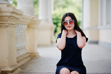 Brunette girl at black dress, sunglasses sitting on stairs of old vintage house, posing at street of city.