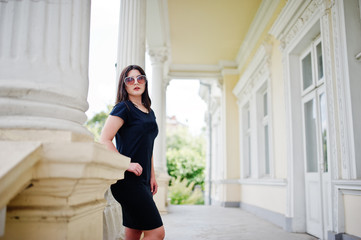 Obraz premium Brunette girl at black dress, sunglasses posed against old vintage house, at street of city.