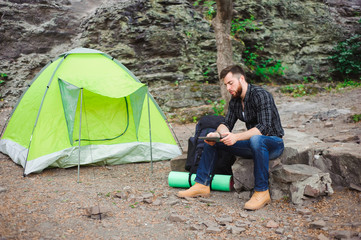 Fototapeta premium Traveler near a tent with a tablet in the evening. Rocky mountains background.