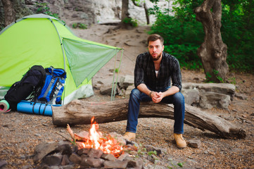 Traveler man relaxing, looking at the fire and dreaming at camping tent outdoors on nature.