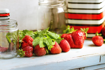 Fresh ripe garden strawberries and melissa herbs with empty glass and vintage metal jars for jam on blue white wooden kitchen table. Rustic style, day light, copy space